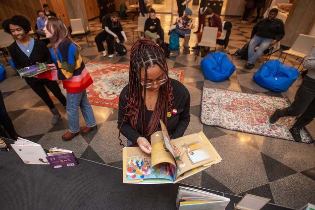 Photo: Sydney Villegas, a young Black woman with burgundy box braids flips through the "Julian is a Mermaid" book from the collection of banned books on display at the stage in front of her. Bean bags, chairs, and rugs are seen behind her facing the stage for storytime. Other students can be seen flipping through other books, sitting in the chairs, and wandering around.