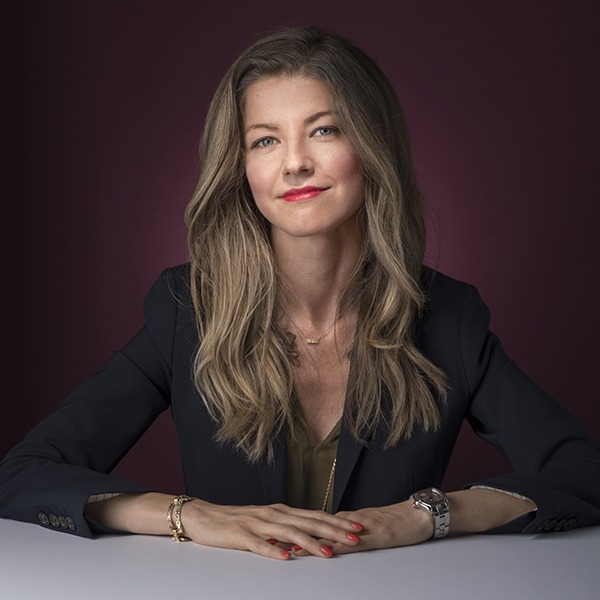 Photo: Headshot of Chiara Longoni. A white woman wearing an olive green blouse and black blazer sits and poses with hands folded on the table in front of her. She smiles in front of a burgundy backdrop.