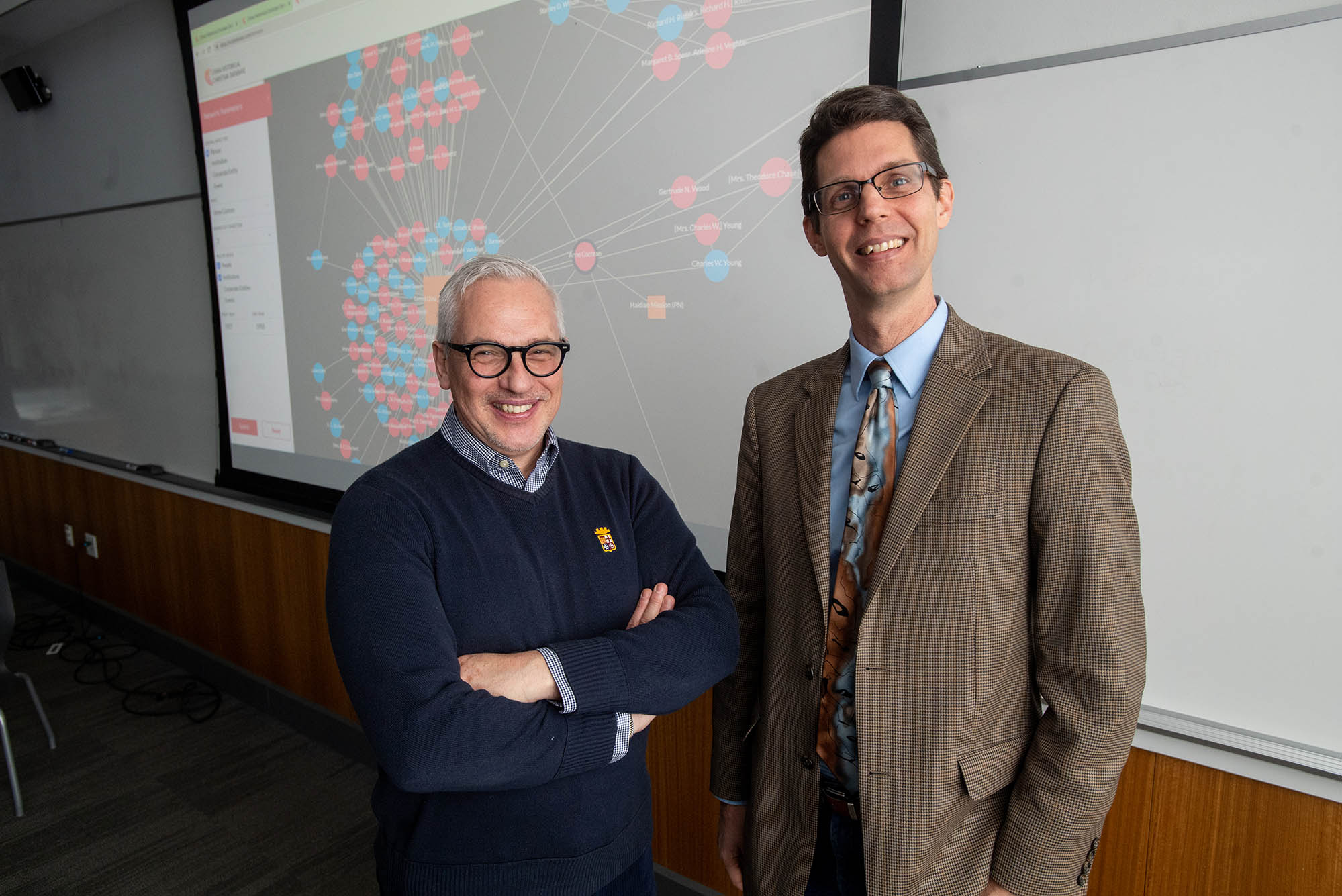 Photo: Eugenio Menegon (left) poses for a photo with Daryl Ireland (right). Menegon, a man with white gray hair and wearing black glasses, a light blue, checkered collared shirt and navy blue sweater smiles and poses with arms crossed. Ireland, a man wearing glasses, light blue collared shirt, gray tie, and brown blazer, smiles and poses with hands in pockets. Both stand in front of a projector displaying a representation of the global network they're working on.