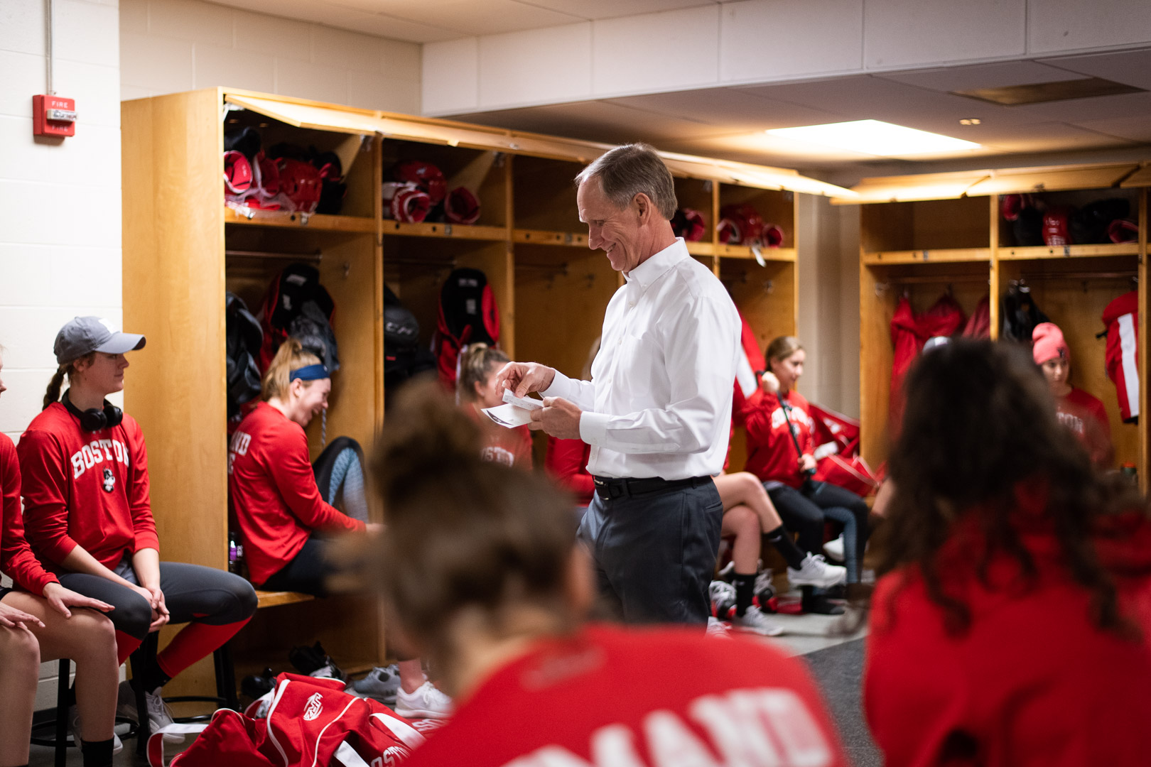 Photo: Brian Durocher a white man wearing a white collared shirt and black pants, smiles as he looks over his notes and speaks to the women's ice hockey team as they sit in their locker room.
