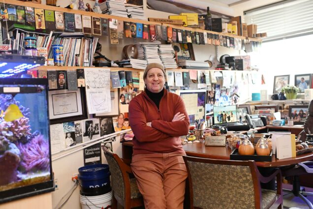 Photo: An Argentinian man with orange slacks, a red sweater with a black turtleneck underneath. He stands in the center of his office. To the left, an aquarium is featured. Above the aquarium tank are shelves of books. Lining the selves are chocolate wrappers from unique chocolate bars. On his right, there is a huge desk with tons of miscellaneous merchandise on top. Some of those items are a yerba mate pot, a silver plaque, and a wooden model of a motorcycle. Two chairs sit in front of his desks. Throughout the room are tons of personal pictures, framed and loose. Mostoslavsky smiles for the photo