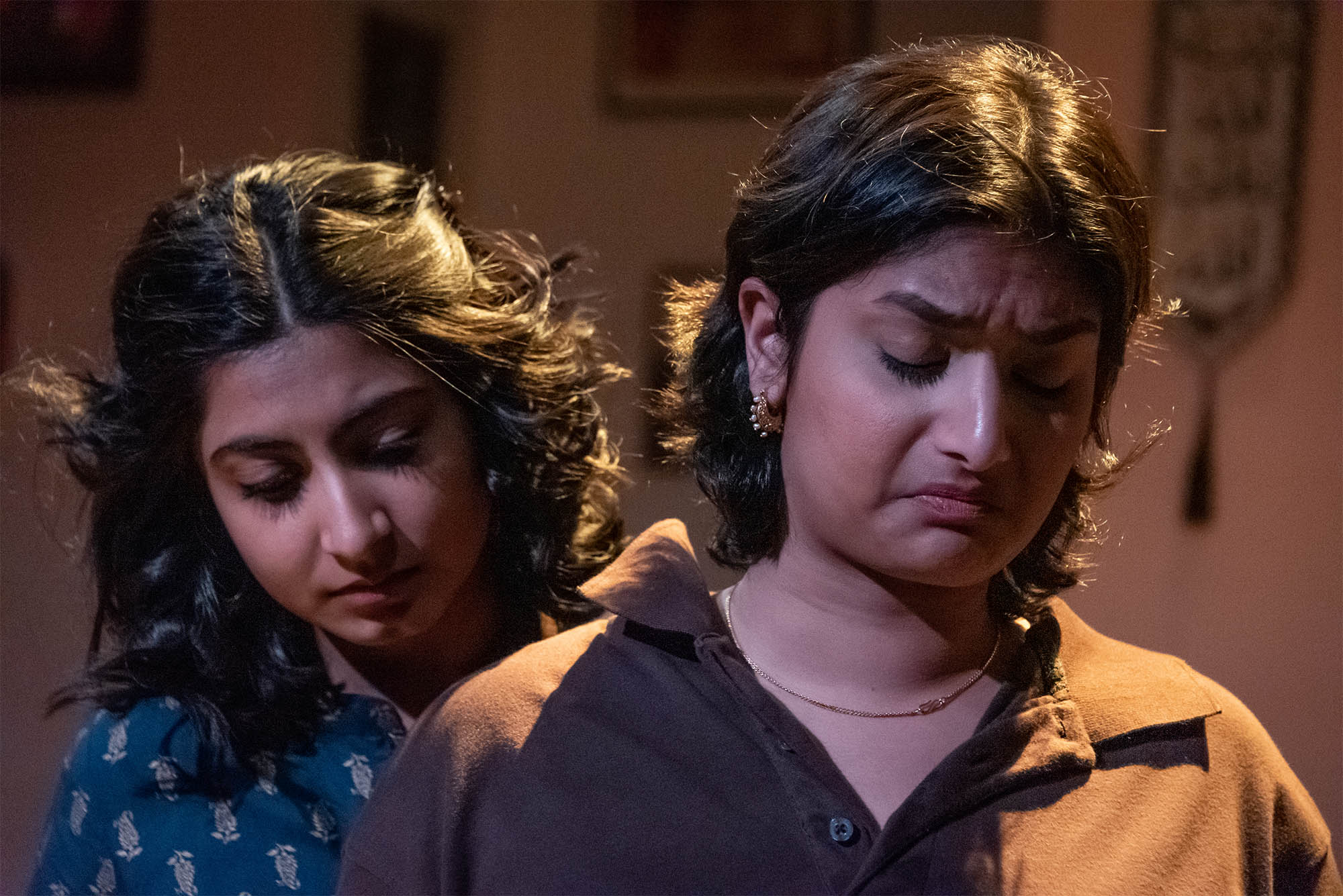 Photo: Sushmita Udoshi (COM’24) as Amal and Vidisha Agarwalla as Mashal in Jado Jehad, a coming-of-age drama set in Pakistan. Two young Brown woman look down and despondent as they act on stage. The woman in the front wears a brown shirt and the other stands behind her, wearing a blue collared shirt. A soft spotlight shines on both.