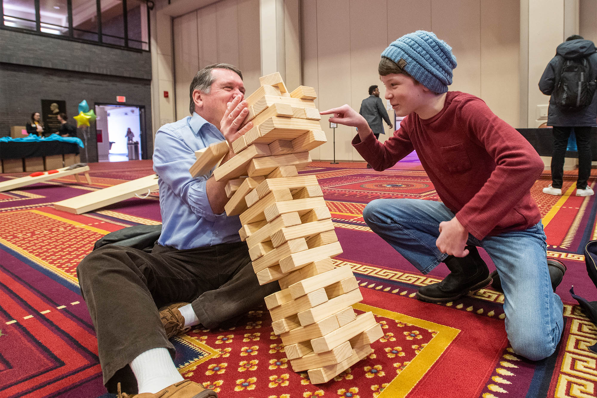 Photo: An older white man wearing a light blue collared shirt and brown pants sits on the carpeted ground next to a young white teen wearing a blue beanie cap, red sweatshirt, and jeans. They both laugh as large Jenga blocks topple towards the older man.