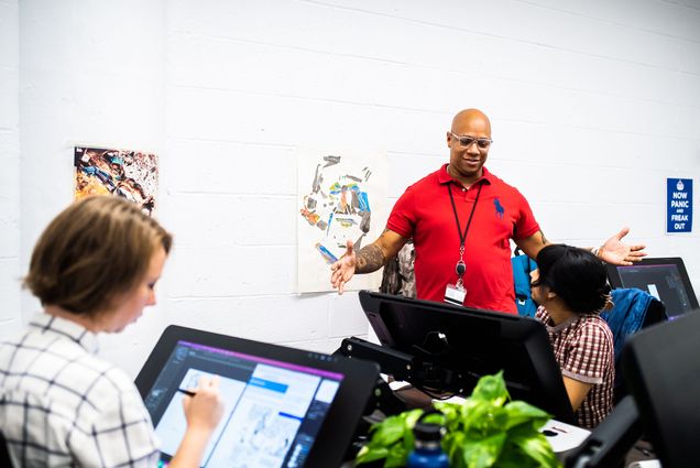 Photo: Joel Christian Gill teaches a new MFA program in Visual Narrative on November 7, 2022. A black man wearing a short-sleeved red collared shirt gesticulates with arms outstretched as he speaks to a student sitting in front of an electric drawing tablet. Other students can be seen in the bright classroom working on similar tablets.