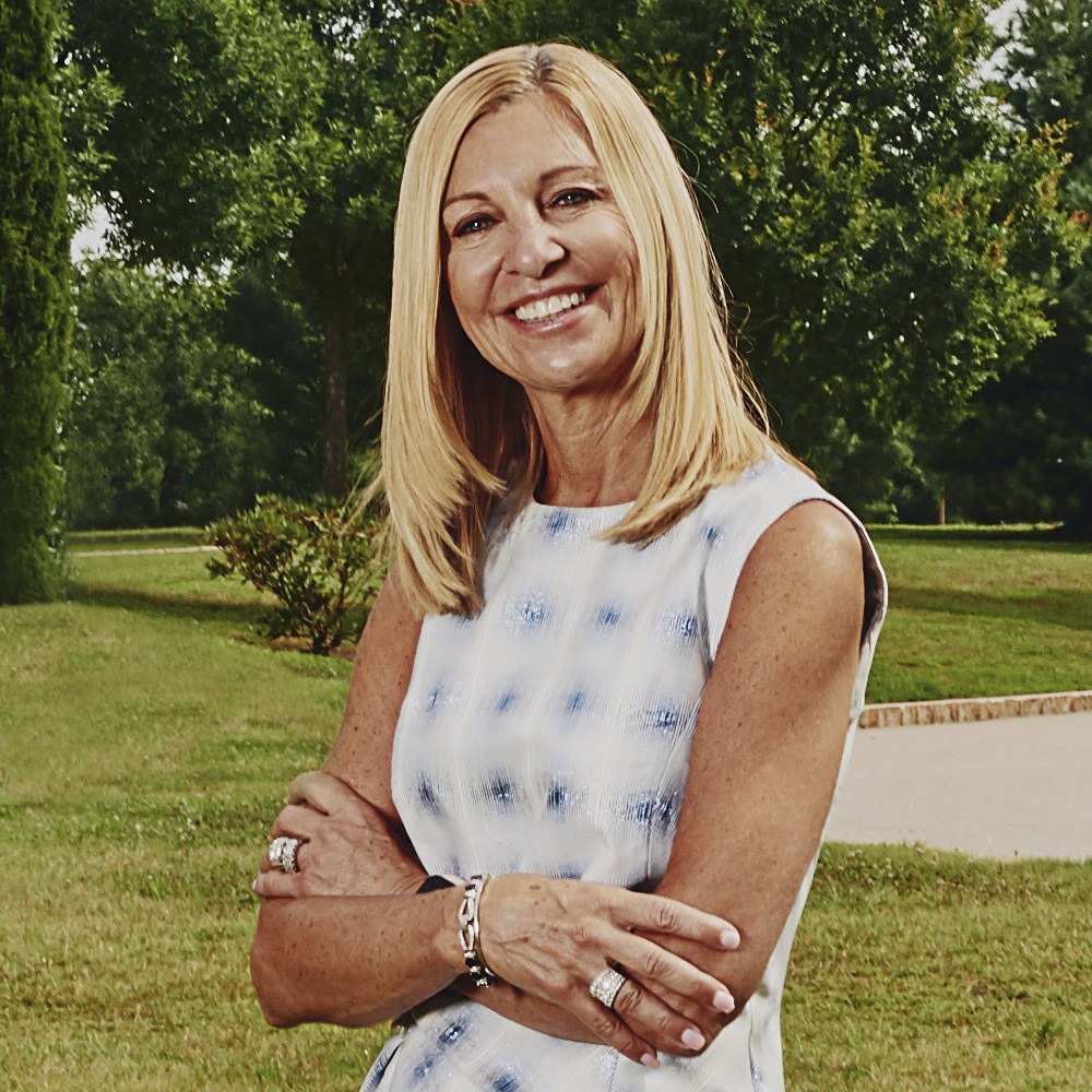 Photo: Karen Lynch in a white and blue patterned dress, her arms crossed, posing in front of a sculpture (cropped out). She stands in a grass field.
