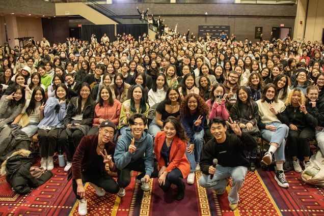 Photo: A large group of mostly Asian students sit in chairs in an audience in a large auditorium-style room, as four people squat and pose in the front. In the front, from left are: Kinh Vũ, assistant professor of music and music education, CFA; Brian Nam, CEO and founder of Dive Studios, the creator of the daily self-care app Mindset; Hyeouk “Chris” Hahm, professor and associate dean of research, School of Social Work; and K-Pop star Eric Nam.