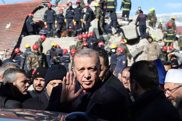 Photo: Turkish President Recep Tayyip Erdogan, an older tan man with grey hair and mustache, tours the site of destroyed buildings during his visit to the city of Kahramanmaras in southeast Turkey, two days after the severe earthquake that hit the region. Rescuers and people can be seen behind him standing and working through large piles of rubble and debris.