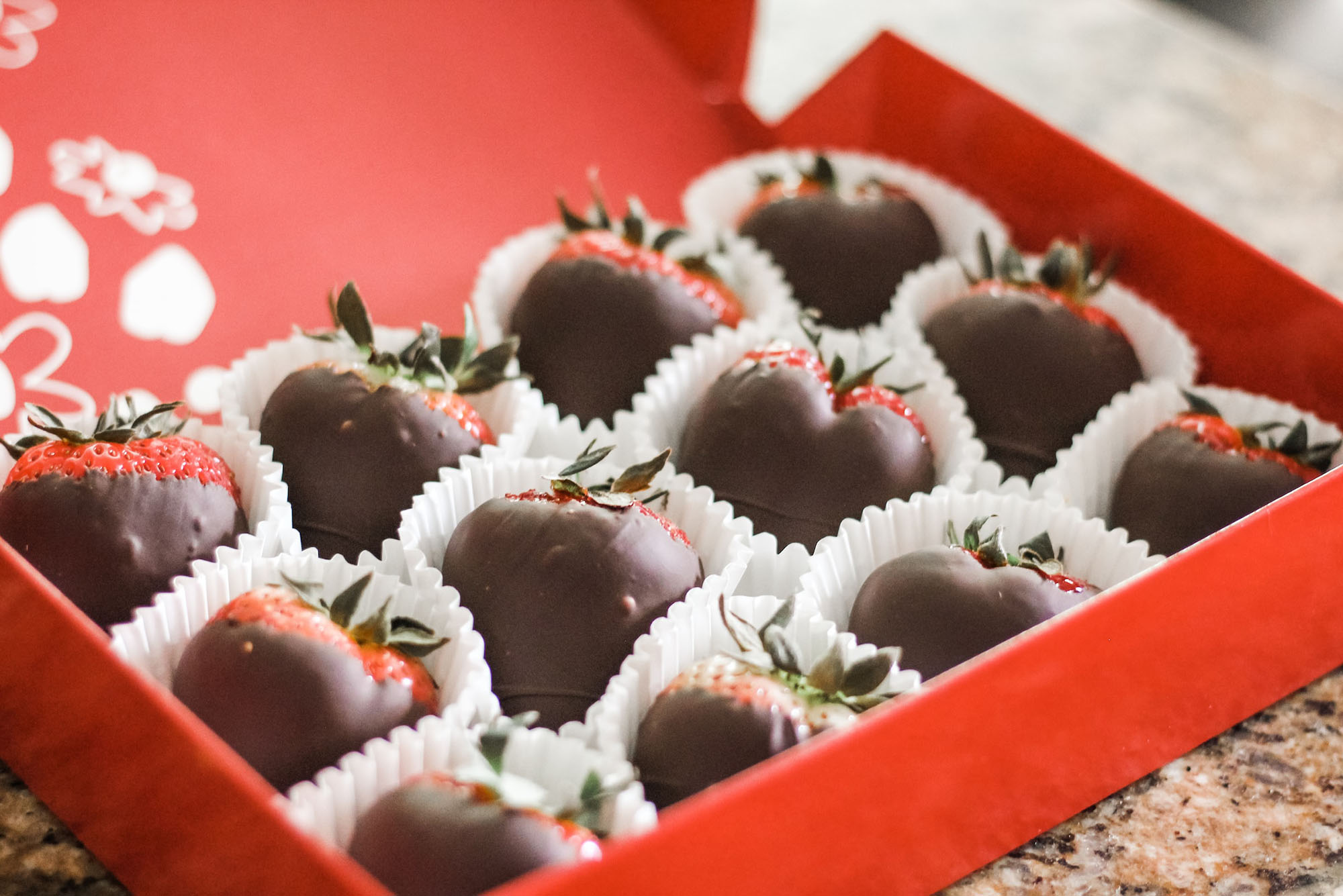 Photo: A red rectangular box of chocolate covered strawberries. The strawberries rest in a small, white paper holders.