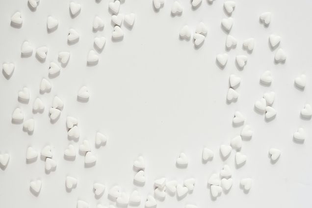 Photo: A bright overhead shot of a white table with scattered white heart-shaped Valentine's candies. Candies are semi-arranged so that there is a large space in the center.