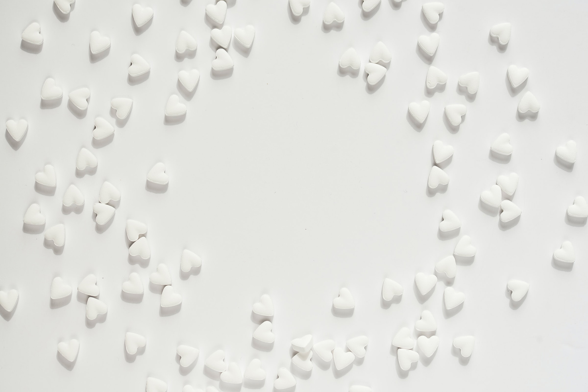 Photo: A bright overhead shot of a white table with scattered white heart-shaped Valentine's candies. Candies are semi-arranged so that there is a large space in the center.