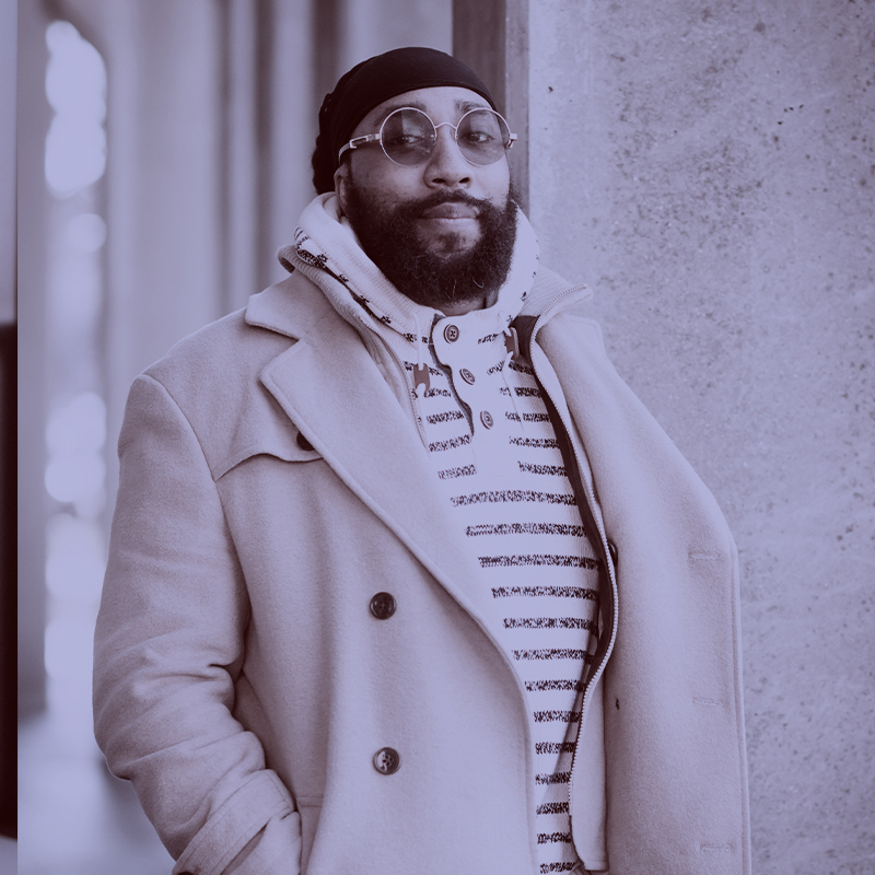 Photo: Dr. Gregory Benoit, a Black man wearing a black headscarf, navy and cream sweater, and tan jacket, poses for a photo with hands in jacket pockets. A light purple filter overlay is placed over the photo.