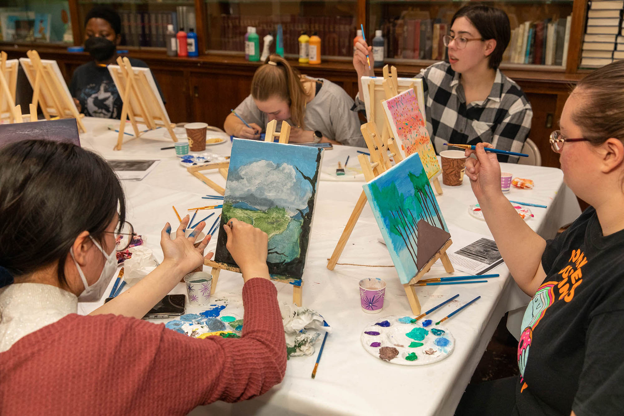 Photo: BU students are painting on mini canvas on a plastic table. Various different painted landscapes are on the canvases with miscellaneous paintbrushes on the table.