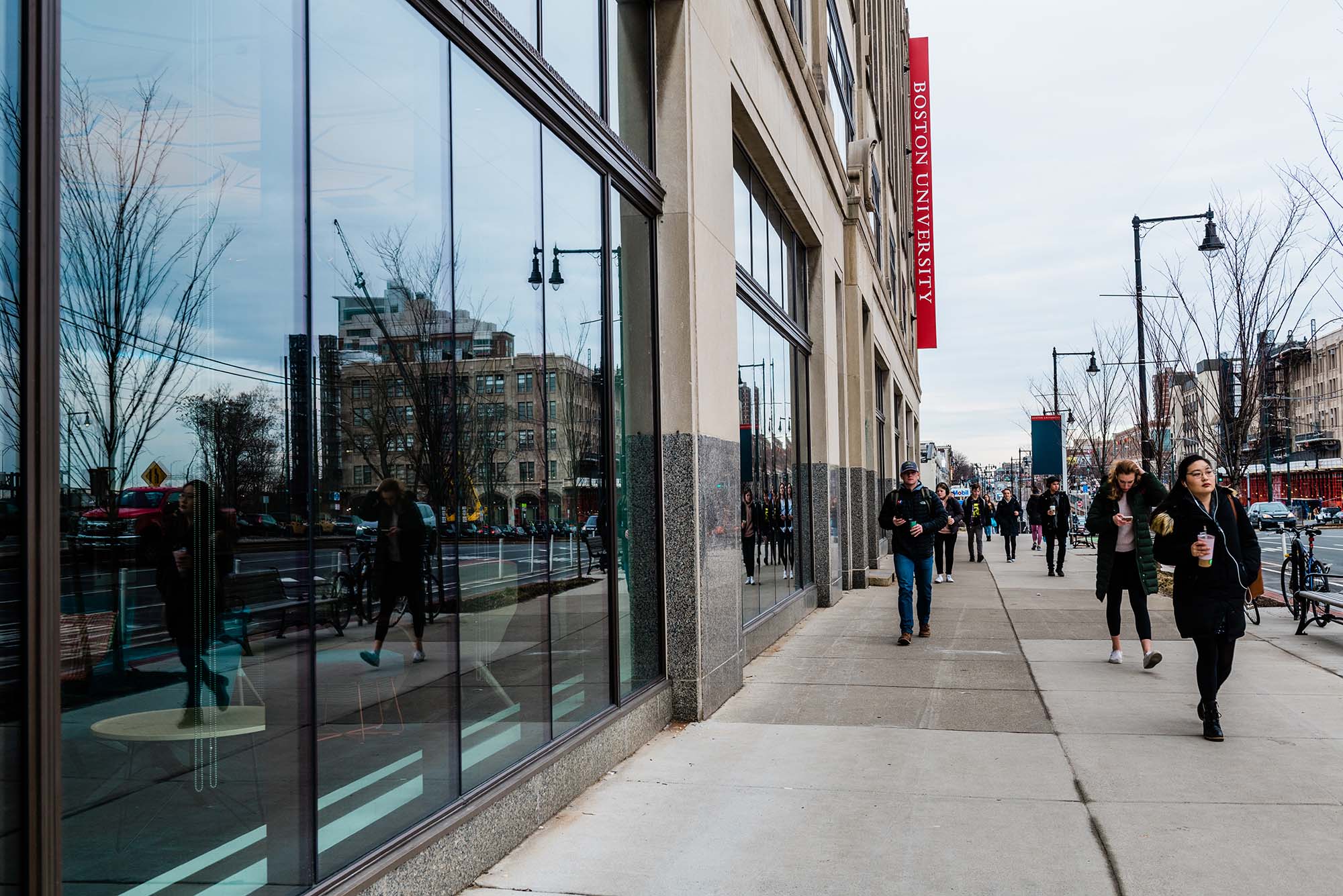 Photo: On the left, a glass wall of a BU building on 808 Commonwealth Ave. There are students walking on the sidewalk on the right. They are wearing winter coats, indicating it's cold outside.