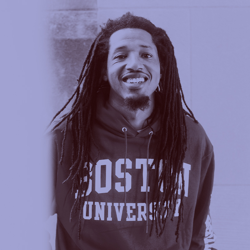 Photo: Darien Pollock, a Black man with long dreadlocs and wearing a "Boston University" hoodie, smiles and poses for the photo. A light purple filter overlay is placed over the photo.
