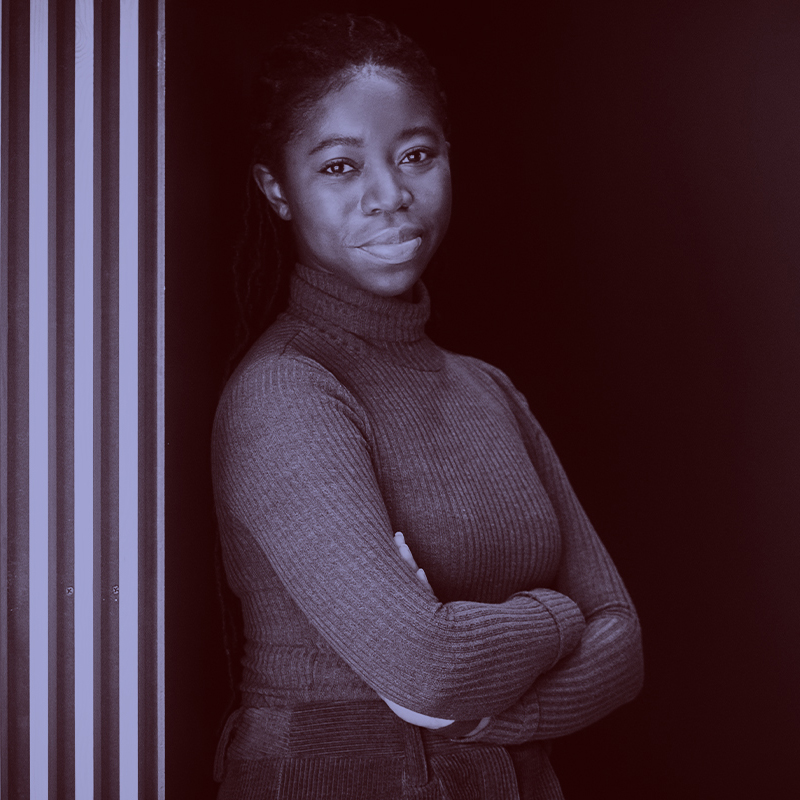 Photo: Tomeka Frieson, a young Black woman wearing a dark gree turtleneck sweater, poses with arms crossed for a photo at CDS. A light purple filter overlay is placed over the photo.