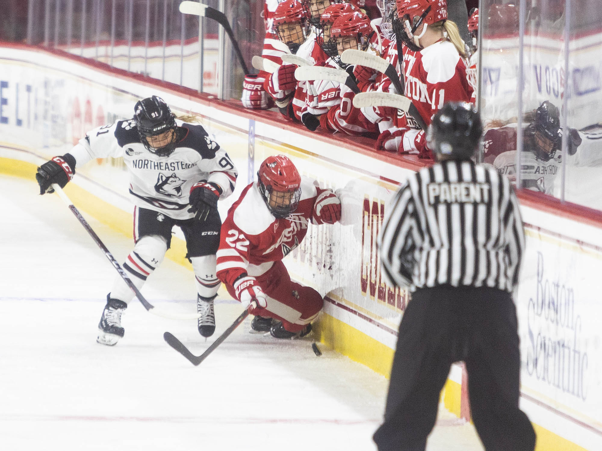 Photo: A NE hockey player in a white and navy uniform checks a BU hockey player against the wall. Behind the wall are the BU hockey team that are currently not on the ice. The BU hockey player is in an all red uniform with white detailing. The referee is in the foreground, bottom right-hand corner of the photo. 