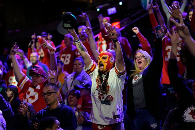 Photo: A crowd of Kansas City Chief fans cheer. The focal fan is in a red and gold mask with a black lanyard and a white jersey. Next to the fan, on the right, is another fan yelling with a black cardigan and a grey t-shirt. She has short gray hair. Everyone around them is wearing a Kansas city jersey. The lighting in the area they are in is purple and blue.