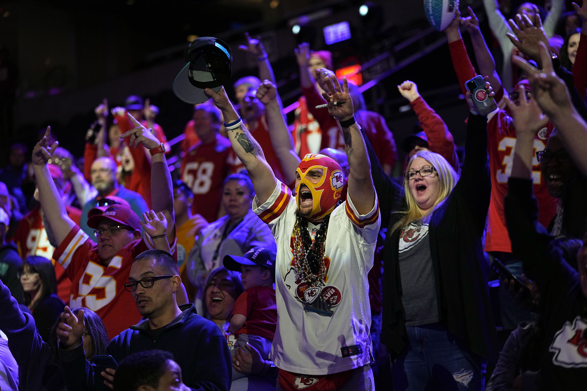 Photo: A crowd of Kansas City Chief fans cheer. The focal fan is in a red and gold mask with a black lanyard and a white jersey. Next to the fan, on the right, is another fan yelling with a black cardigan and a grey t-shirt. She has short gray hair. Everyone around them is wearing a Kansas city jersey. The lighting in the area they are in is purple and blue.