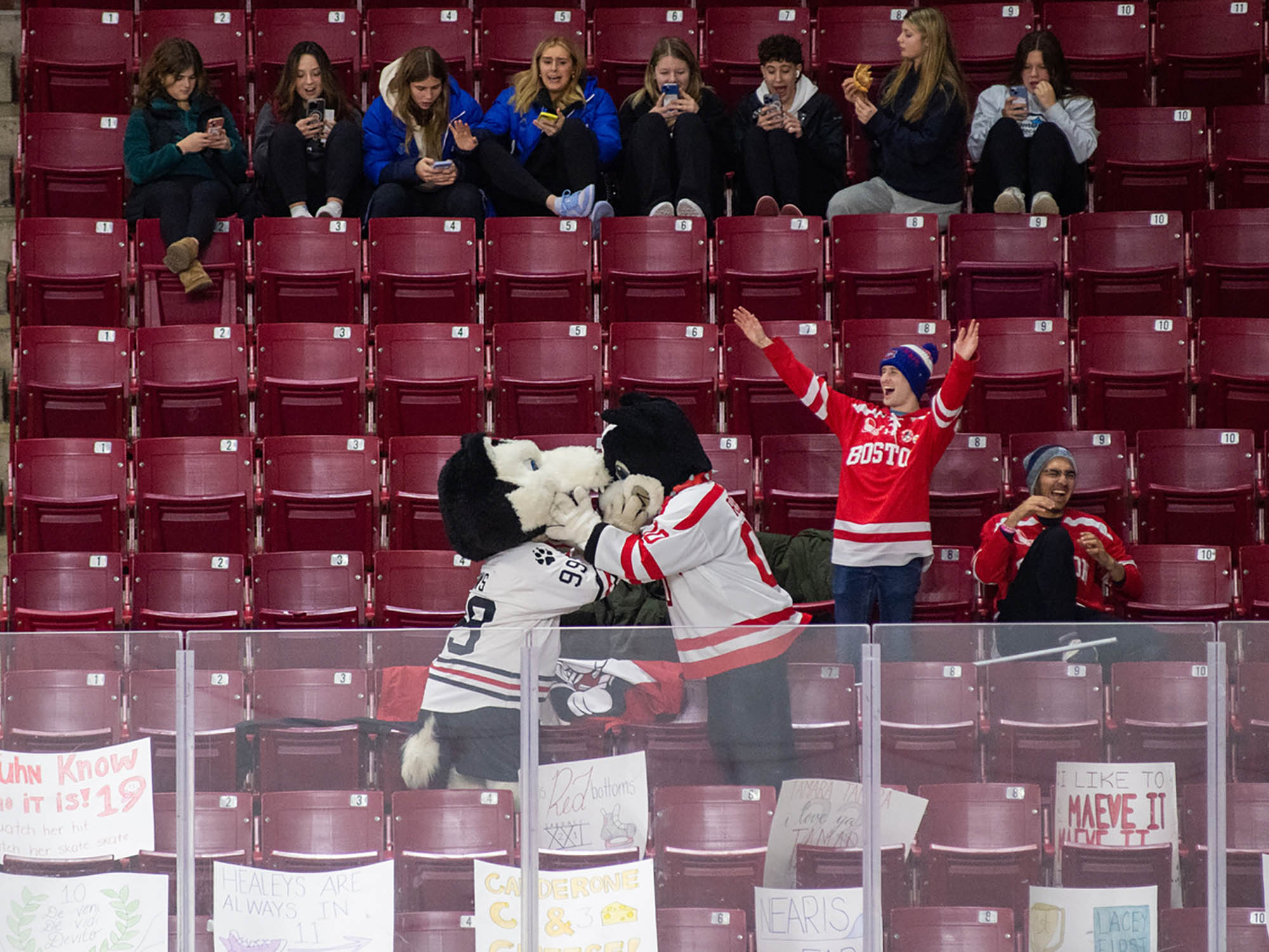 Photo: Two mascots, BU Terrier and NE Husky, fake fight in ice rink hockey stands. The chairs around them are a dark red. To the right of the mascots, a BU fan cheers with a red jersey and a black beanie on. He sits next to another individual with a red letterman jacket and a grey and red beanie. Above them, five rows up, are a group of feminine-presenting individuals with sports-wear on. They are talking amongst themsevles.