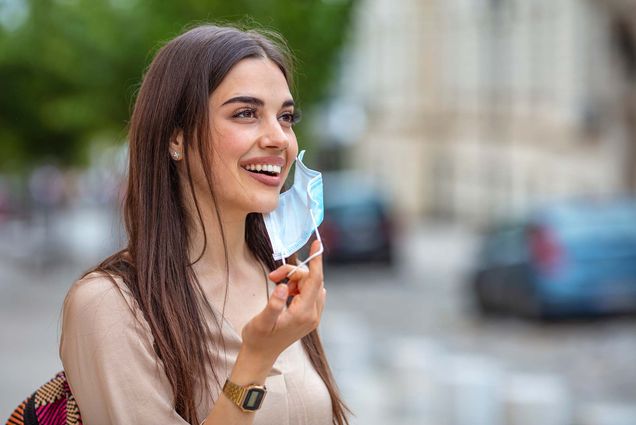 photo of a young, light-skinned woman with brown hair taking of her blue facemask and smiling. she wears a backpack and a tan blouse. a street is seen blurred behind her in the background.