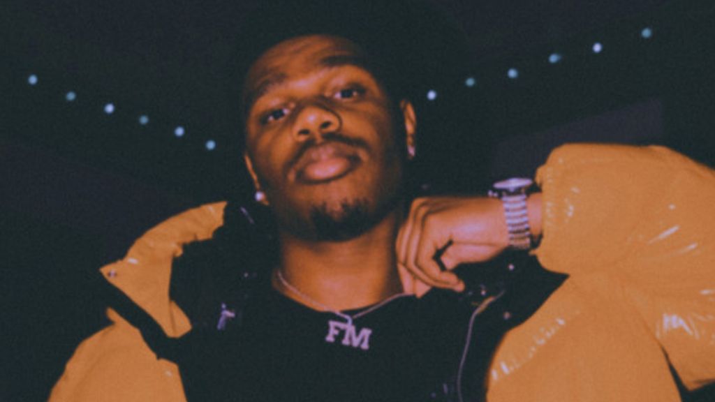 Headshot of musician Fridayy sporting a yellow-orange puffer coat with a black shirt and a necklace with the letters "FM."