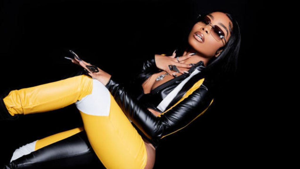 Photo of musician Lola Brooke in a tight yellow and black leather suit in front of a black background.