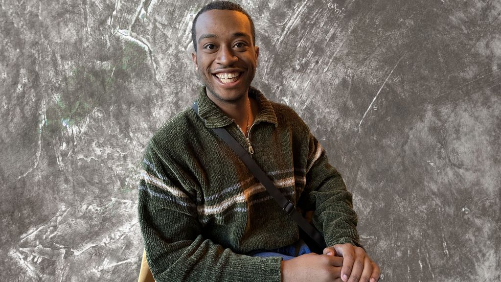 The headshot of musician Rozime Lindsey in a green sweater with a brown and white stripe is in front of a stonewashed background.