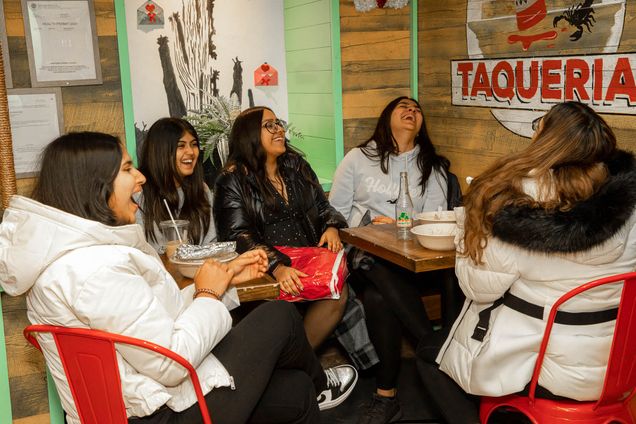 Photo: Five young feminine-presenting individuals laugh and dine at El Jefe's Taqueria. They sit at two, parallel tables. From left, the individual wears a white jacket and long black hair. Next, the individual has a light, gray shirt and long black hair. The next individual has square glasses, a black button up, and long black hair. They are throwing their head back to laugh. Second to last, on the right, the individual has a light gray hoodie on and long, black hair. The right-most individual has a white puffer coat on with black fur trim on the hood. Their hair is long, with black roots and bleached from the crown of their head, down.