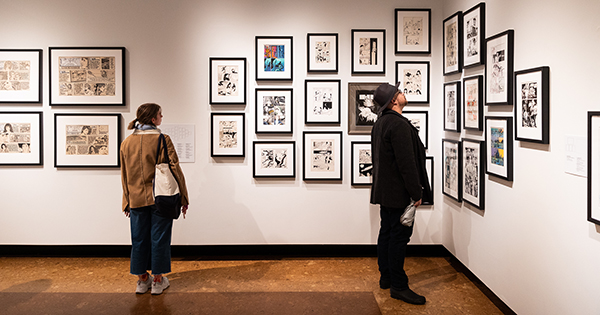 Photo: Two people are looking at framed artwork and comic pages in a gallery. The individual on the left is wearing a tan jacket, a white canvas bag, denim pants, sneakers, and a scarf in their hair. The individual on the right is wearing an all black ensemble with a black fedora.