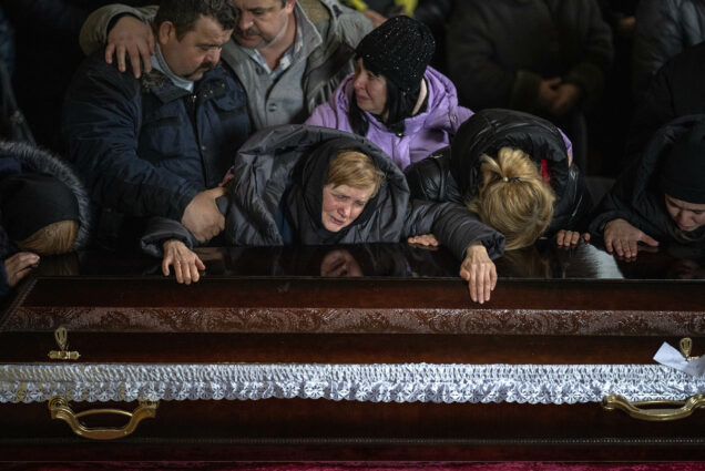 Photo: A Ukrainian woman and her family members gather over a closed casket at a funeral for a Ukrainian solider who died during the Russian occupation of Ukraine. They are wearing heavy outwear indicating it is cold.