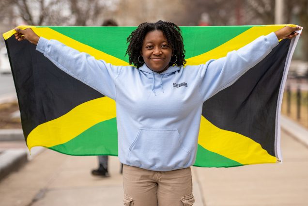 Photo: Brianna Bourne. a Black young woman with a kinky curly twist out smiles as she holds up a Jamaican flag. She wears a light blue hooded sweatshirt and tan pants as she holds up the flag behind her.