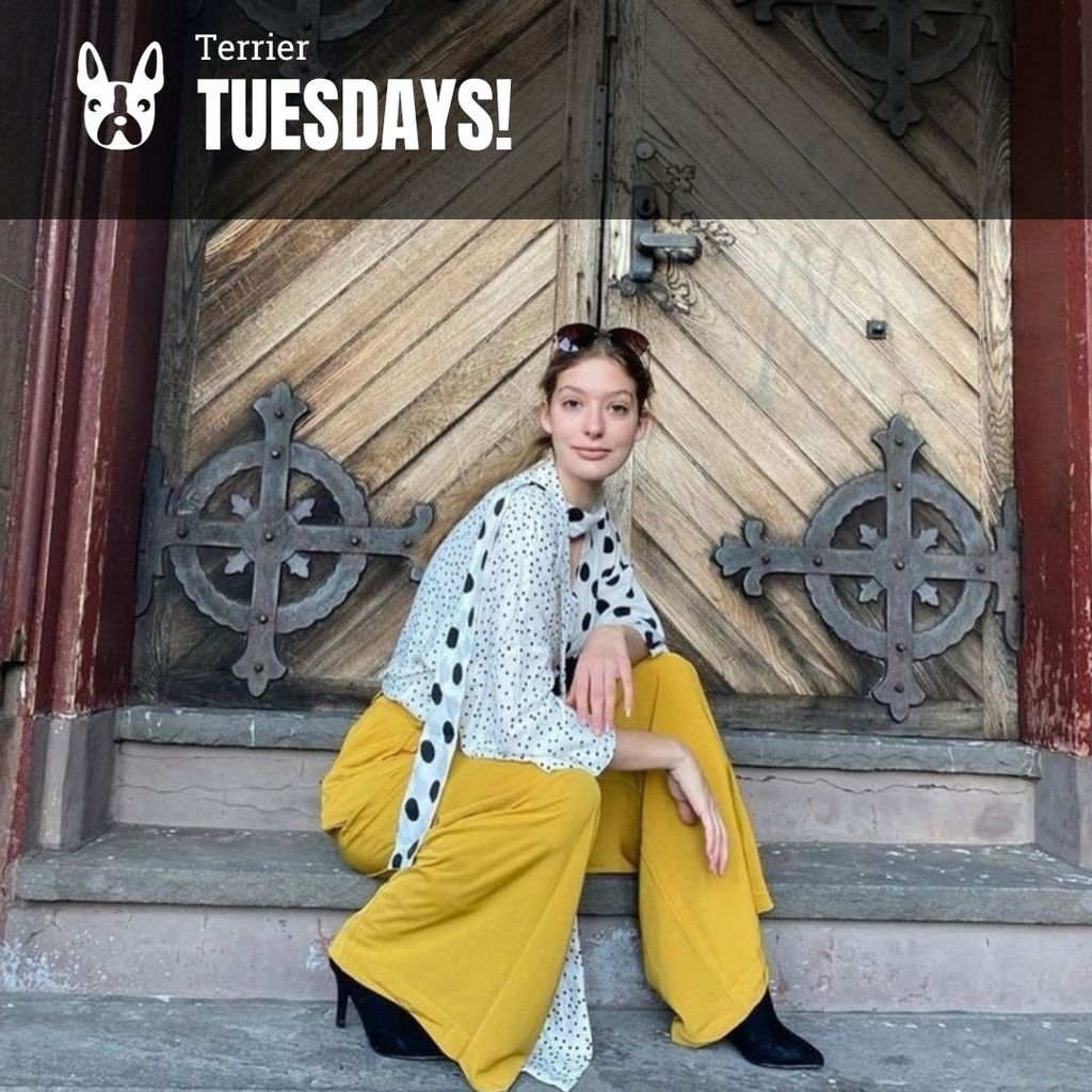 Photo: A white woman sits on the steps of a building with a grand door behind her. She wears yellow pants and a stripped blue shirt. She is looking fiercely into the camera.