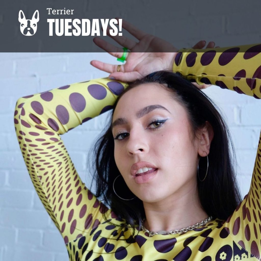 Image: Photo of a young woman posing with arms overhead. She wears a yellow long sleeved shirt with black dots and looks tot he camera. Overlay reads "Terrier Tuesdays!"