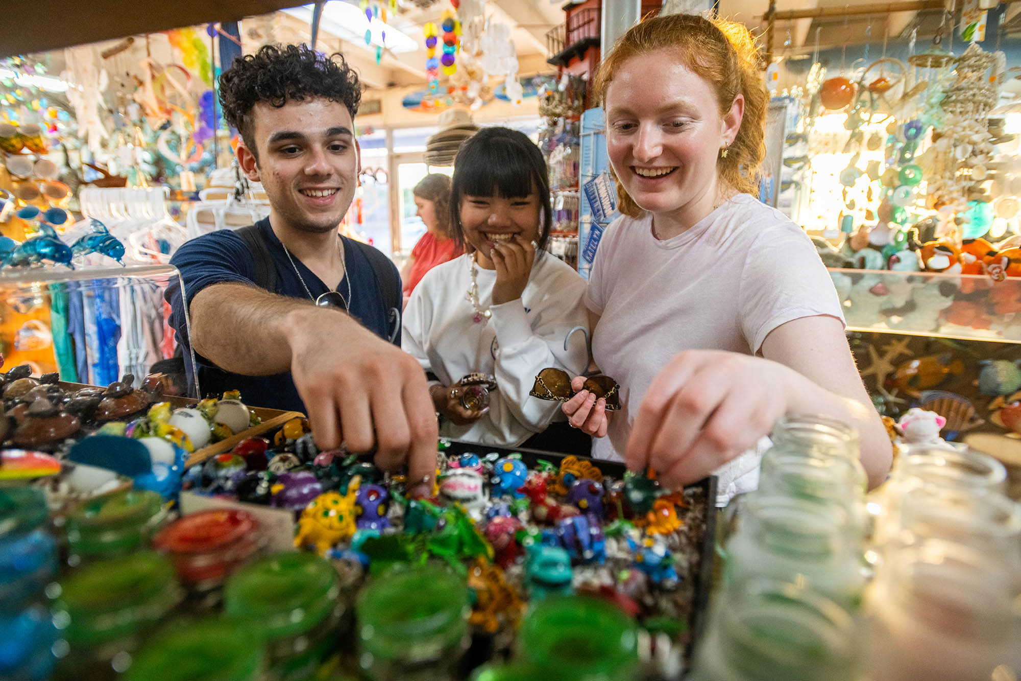 Photo: Three young adults smile as they reach forward and look through glass trinkets in a brightly lit and crowded trinkets shop.