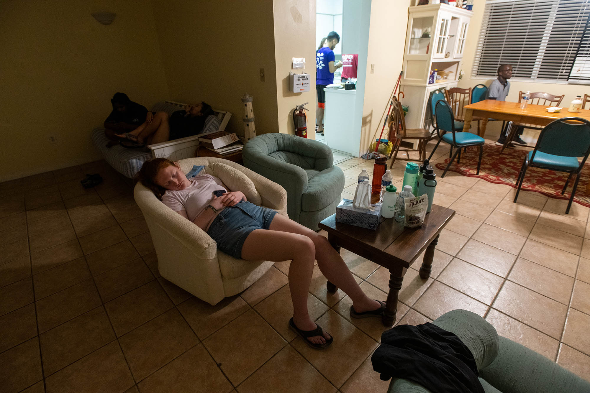 Photo: A young white woman naps in a beige arm chair in a darkened room. In the background, a lit kitchen with another young adult wearing a purple shirt is seen.