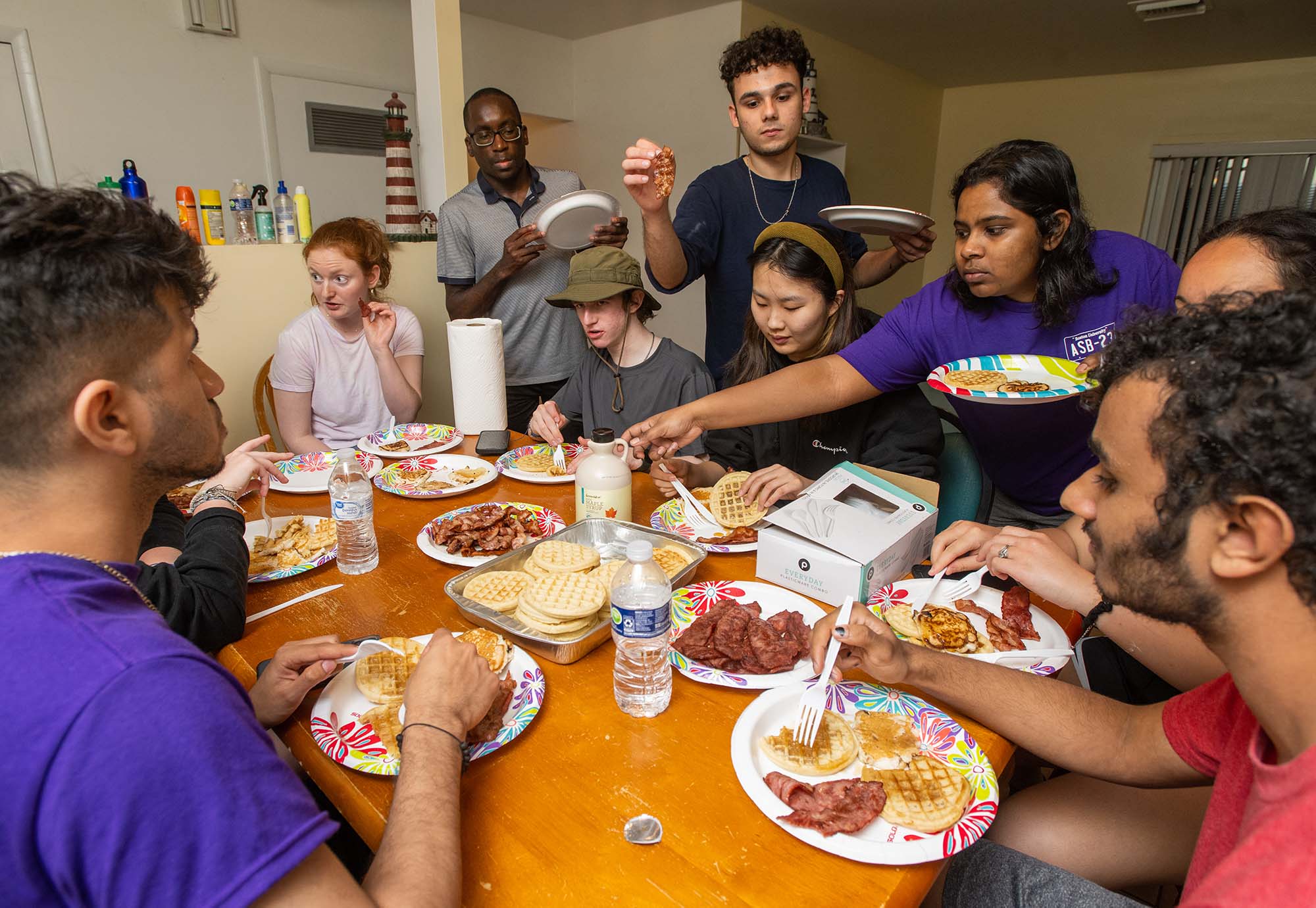 Photo: a group of diverse young adults sit and stand around a large wooden table filled with plates of chicken and waffles. The students serve themselves food, as they chat and laugh.