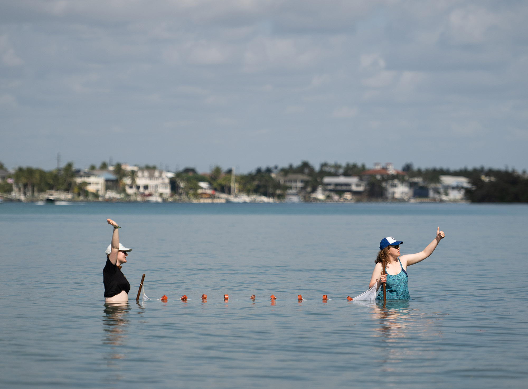Photo: Two young white people stand waist deep in a large body of water and hold up seine netting with one hand as they raise their other arms.
