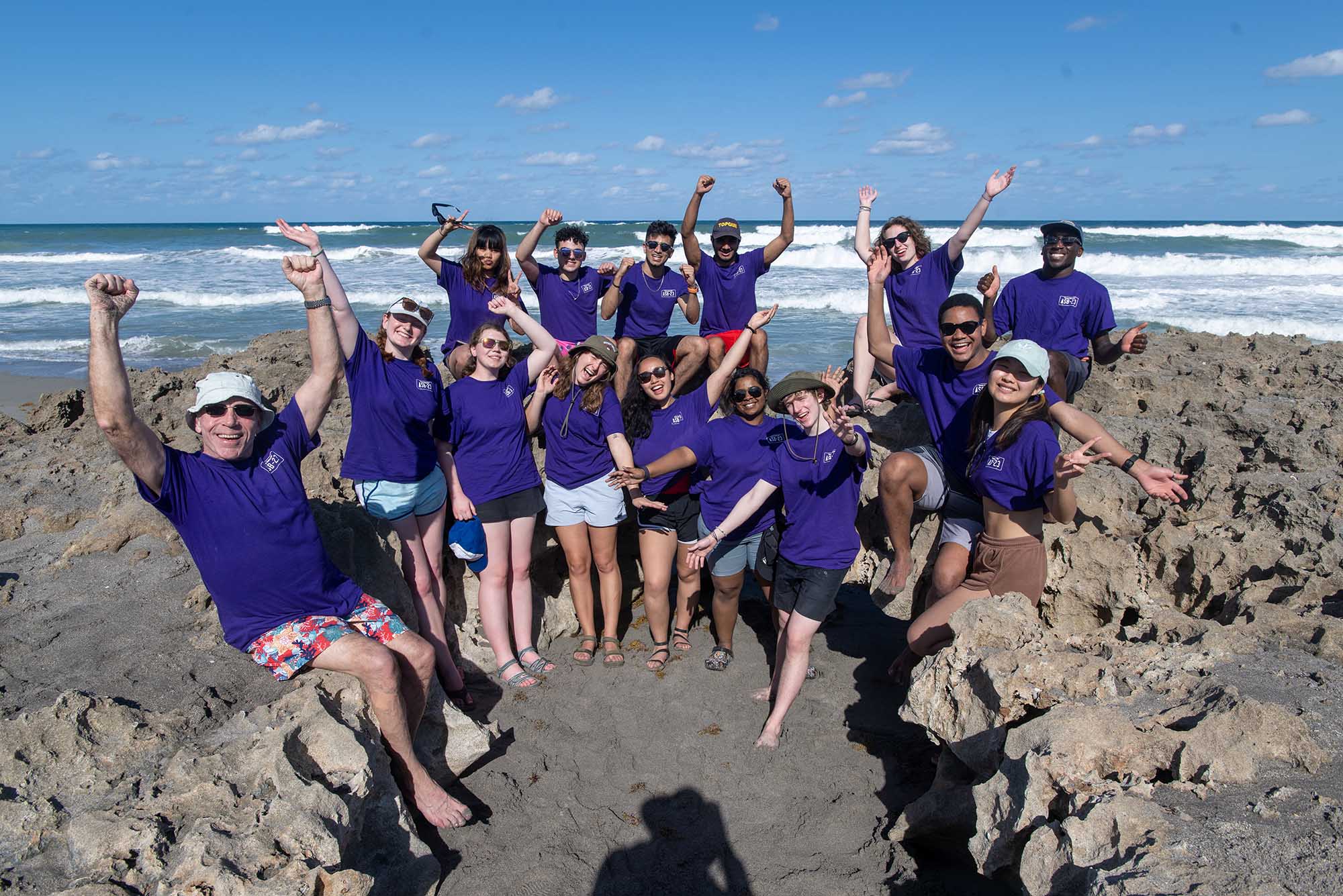 Photo: Group photo a 14 students and one older chaperone. They all wear purple short-sleeved shirts and a variety of hats as they cheer and laugh. They sit and stand around a group of sedimentary rocks on a sandy beach in front of a large body of water on a sunny day.