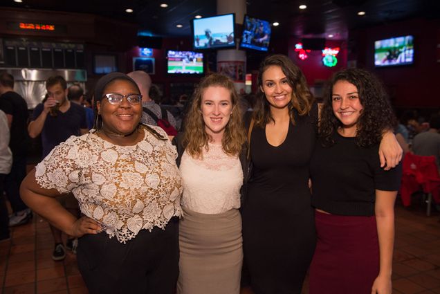 Photo: Four women pose with arms over each other's shoulders for a group photo. (From left) A black woman wearing wearing glasses, a lace white blouse, and black pants, a white woman wearing a cream blouse and tan skirt, a tan woman wearing a black dress, and a tan woman wearing a black short sleeved shirt and burgundy skirt, all pose for a photo together.