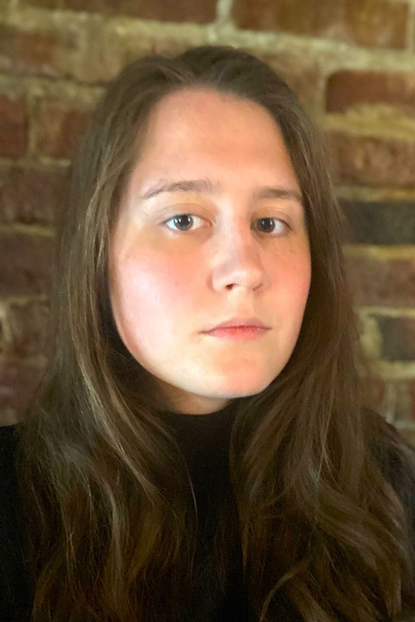 Photo: Headshot of a white-passing woman, she has straight, long, brown hair, rosy cheeks, and brown eyes. She stares squarely into the camera in front of a brick background.