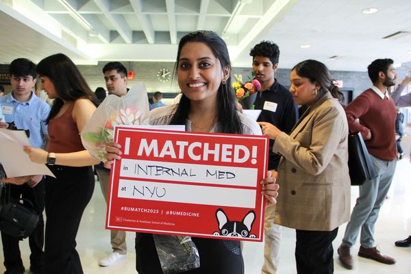 Photo: A young South Asian woman holds up a red sign that reads "I Matched! In Internal Med at NYU". Other people holding similarly styled signs gather and congratulate each other behind her.