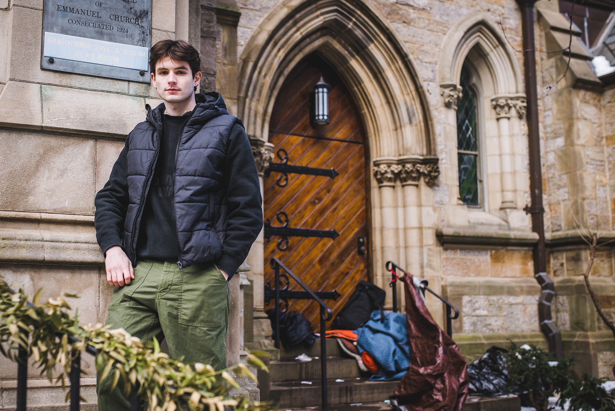 Photo: Food for Thought founder Ben Myers (CGS’22, CAS’24) outside Emmanuel Church, a day shelter on Newbury Street where he films his videos. A young white man with dark brown hair stands with hands in pockets in front of a church with large wooden doors. He wears a black hooded sweatshirt, dark grey puffer vest, and olive green cargo pants. An unhoused person's belongings can be seen scattered in front of the entrance of the church.