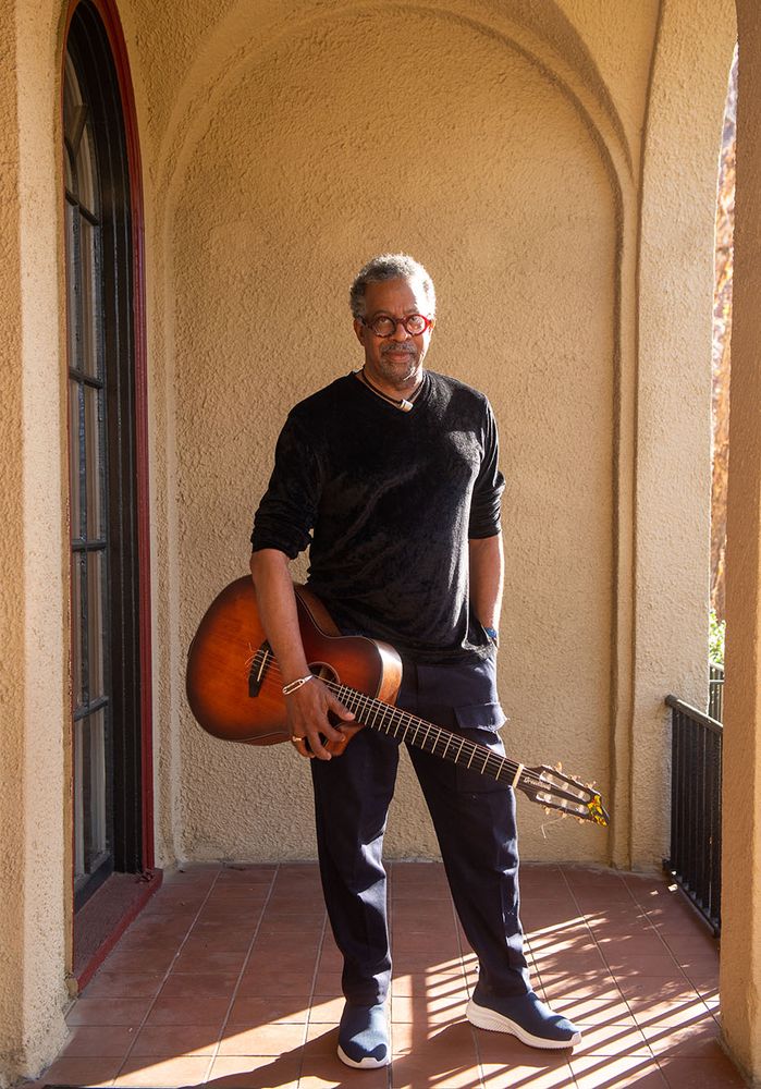 Photo: Bill Banfield, A Black man short graying hair wearing red glasses, a black long sleeved shirt with sleeves rolled up to his elbows, and black pants, poses as he holds his guitar under one arm. He stands at the exterior of a Spanish style building deck as the sun shines on him.