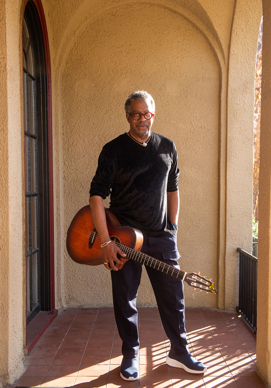 Photo: Bill Banfield, A Black man short graying hair wearing red glasses, a black long sleeved shirt with sleeves rolled up to his elbows, and black pants, poses as he holds his guitar under one arm. He stands at the exterior of a Spanish style building deck as the sun shines on him.