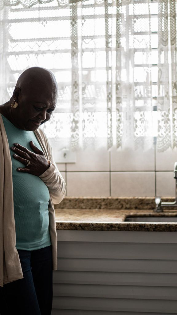 Photo: A senor, Black woman wearing a light blue shirt, tan cardigan, and black pants, has a look of pain on her face as she clutches her chest in her kitchen.