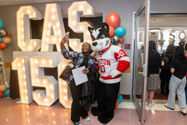 Photo: A young Black woman wearing a black and white plaid collared shirt and black pants takes a photo with a large, Boston Terrier mascot dressed in a BU hockey jersey. A large sign behind them is light with bulbs and reads "CAS 150". Students and others to the left enter a room decorated for a celebration.