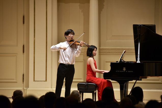 Photo: A young Asian man wearng a white collared shirt tucked into black pants plays a violin passionately on stage. Behind him and to the right, a young Asian woman wearing a long red dress, plays a black grand piano. The silhouettes of the audience can be seen in front them watching.