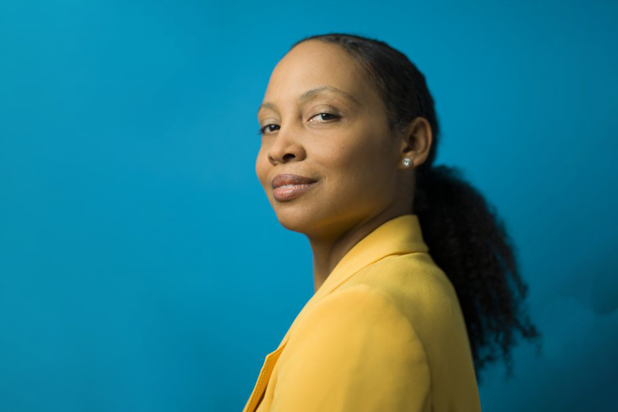 Photo: Esther Ohrt, a Black woman with a slicked back, curly ponytail and wearing a yellow blazer, smiles and poses with a look over shoulder towards the camera. she stands in front of a teal blue background.