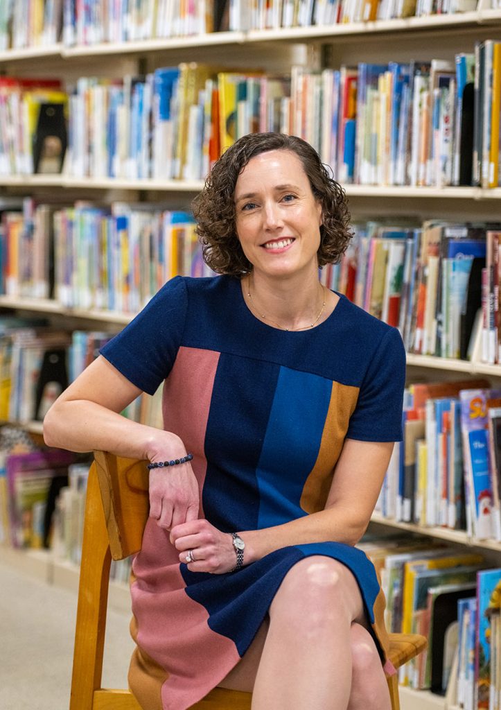 Photo: Portrait of Katherine Frankel. A white woman with short, curly brown hair and wearing a navy blue, blue, and dark pink, and muted orange block-striped dress sits and poses with legs crossed in a wooden chair. She smiles for the camera as she sits amongst bookshelves filled with books.
