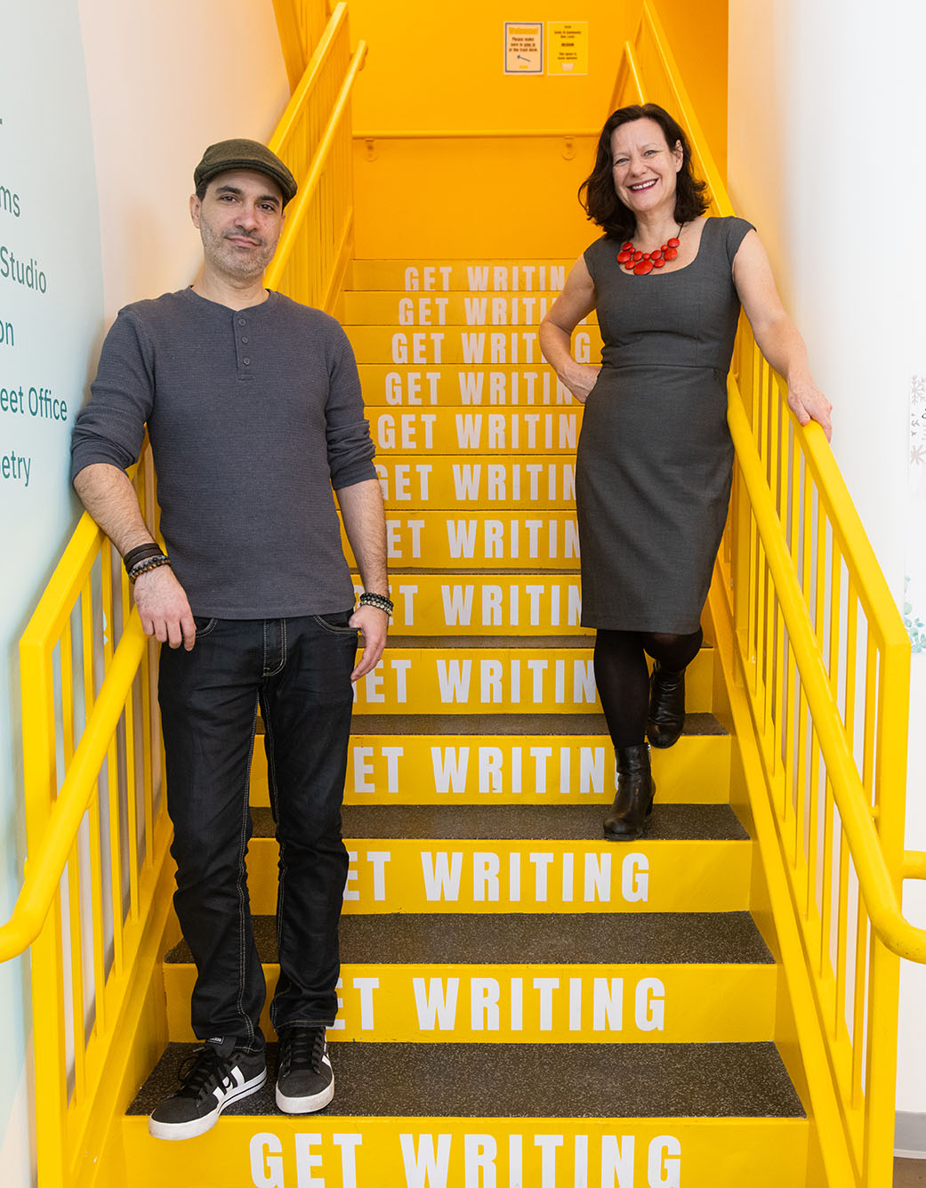 Photo: Artistic director Dariel Suarez (GRS’12) (left) and founder and executive director Eve Bridburg (GRS’97) stand and pose on a set of bright yellow stairs. Each step has the words "Get writing" printed on them in white. A latino man wearing a grey beret, grey shirt, and black pants stands with one arm resting on the railing to his left and smiles. To his right, a white woman wearing a grey dress, black pantyhose, and red chunky necklace smiles and stands with one arm resting and poised on the railing to her right.